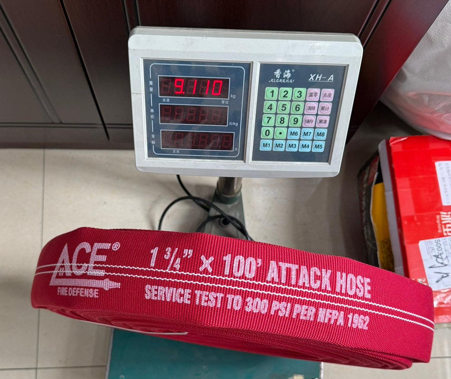 A red ACE fire lifter attack hose laid across a digital scale, with the scale displaying 3710. The setting appears to be a home or warehouse floor with dark wood cabinets, a plastic bag, and a red box in the background.