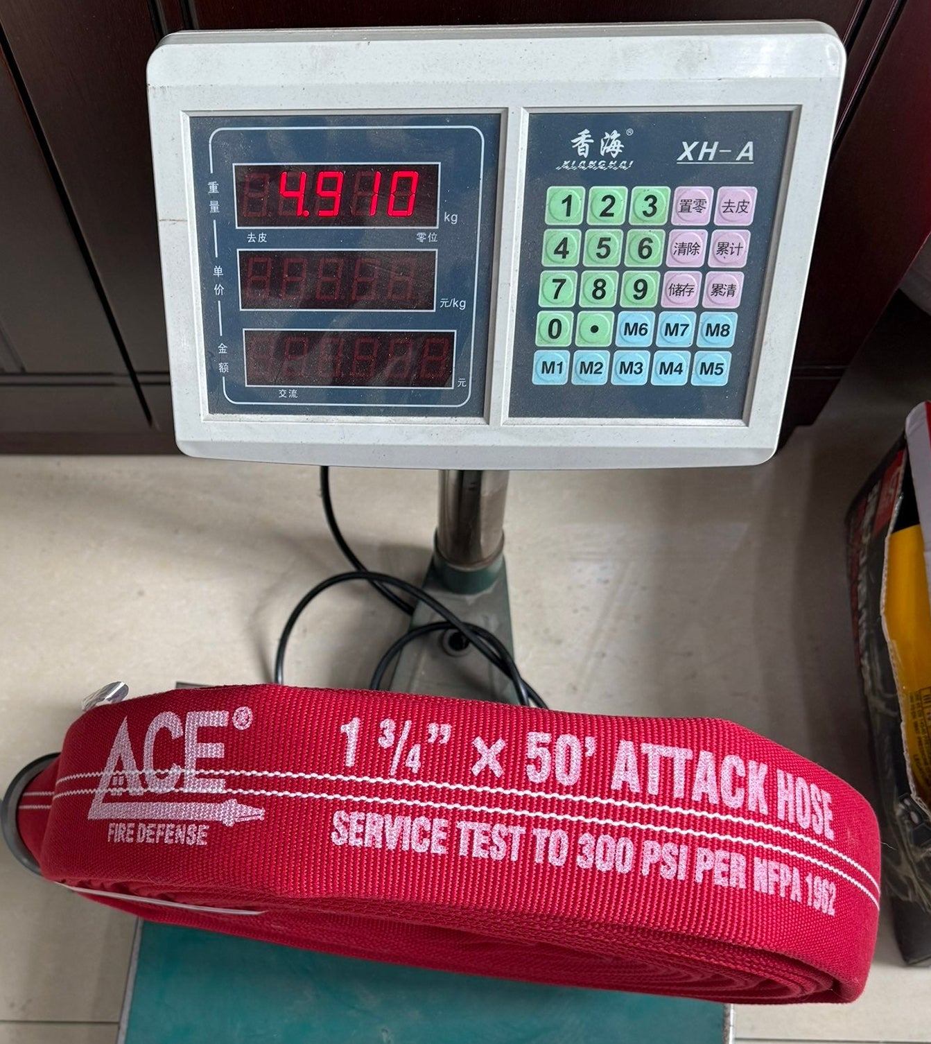 A red fire department attack hose with white text reading 'ACE' and '1 1/4" x 50" ATTACK MRE' resting on a digital scale, with a person's shoes visible at the bottom edge.